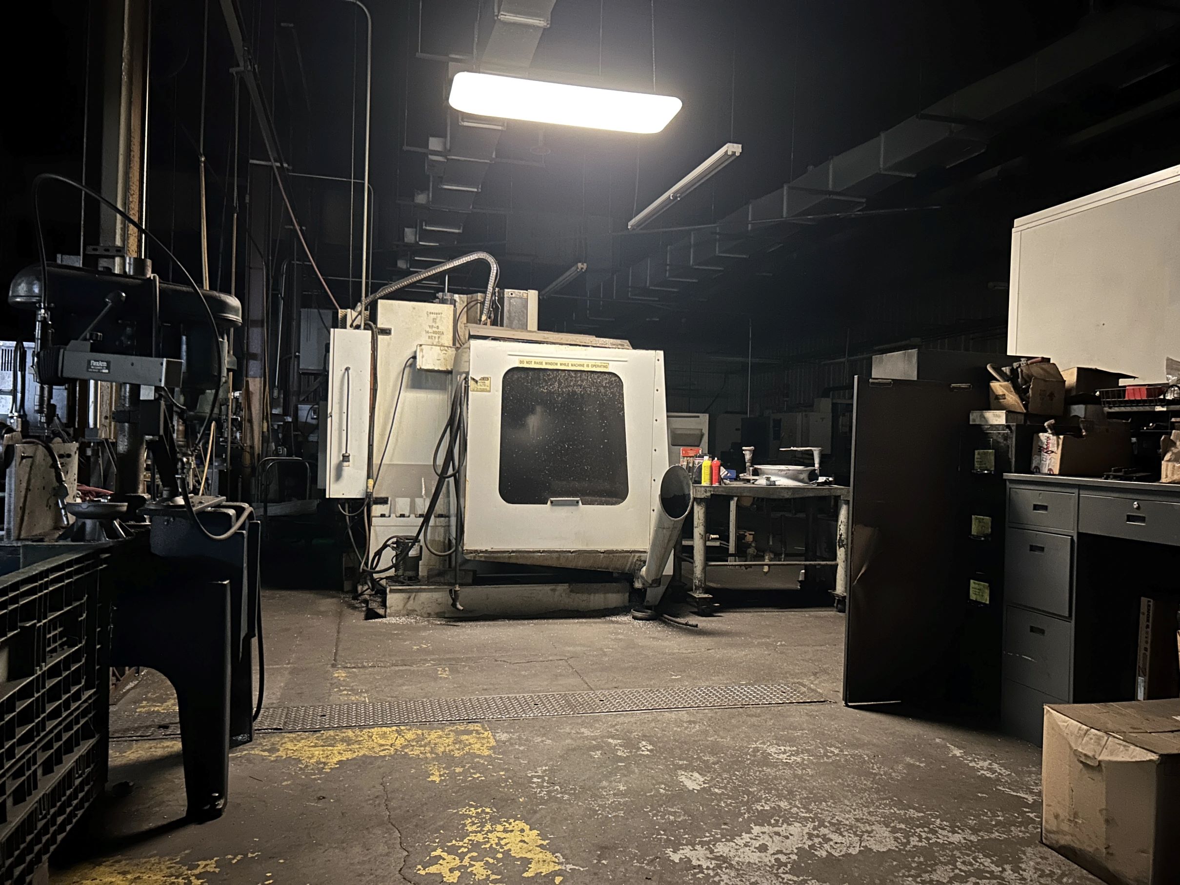 Image of a dirty, white machine in the middle of a concrete floor. The room is dimly lit. The machine is surrounded by other nondescript heavy machinery.